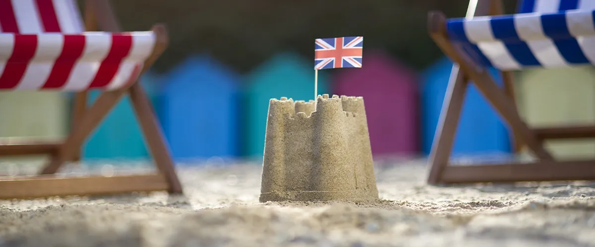 Château de sable surmonté d'un drapeau britannique sur une plage avec transats colorés évoquant les séjours linguistiques en Angleterre