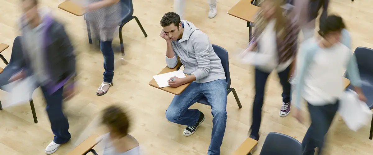 Un lycéen isolé et pensif reste assis à son bureau tandis que d'autres élèves s'agitent autour de lui dans une salle de classe