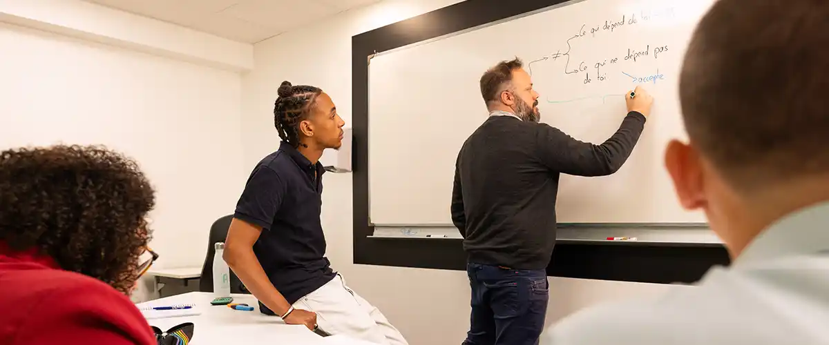 Un enseignant Acadomia écrit au tableau blanc pendant qu'un petit groupe d'élèves attentifs suit le cours dans une salle de classe - photo tous droits réservés