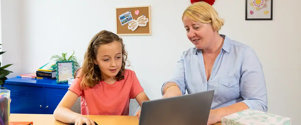 Une élève de primaire concentrée travaille sur un ordinateur portable accompagnée d'une enseignante Acadomia à domicile - photo tous droits réservés