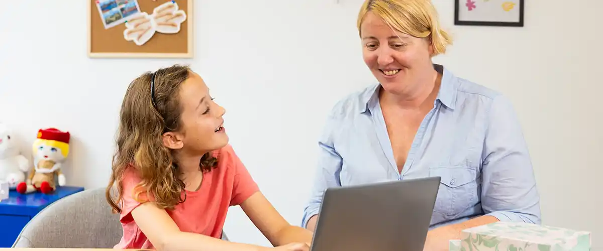 Une écolière souriante partage un moment complice avec son enseignante Acadomia devant un ordinateur portable lors d'un cours à domicile - photo tous droits réservés