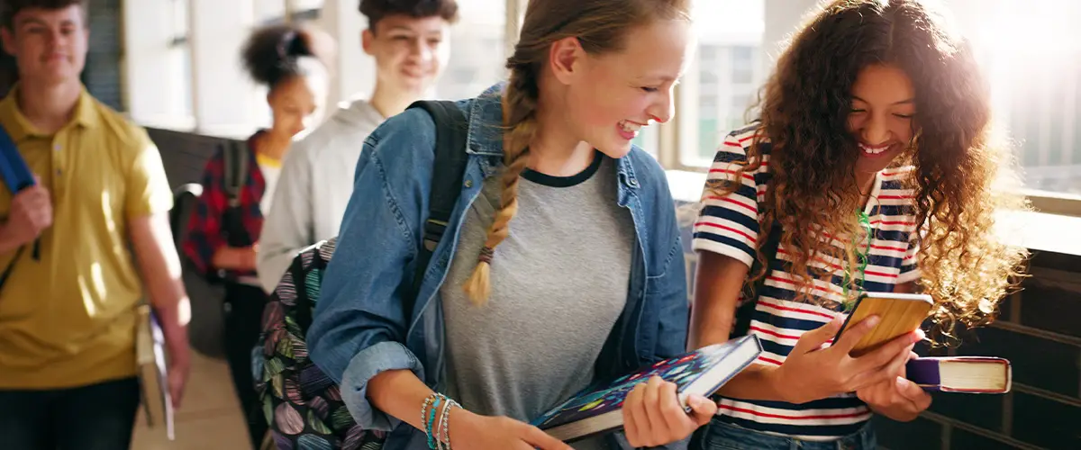 Deux collégiennes souriantes partagent un moment complice en regardant un smartphone dans un couloir de leur établissement scolaire