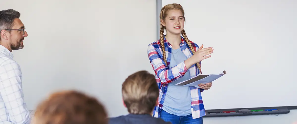 Une lycéenne confiante présente un exposé devant sa classe sous le regard attentif de son enseignant dans une salle Acadomia