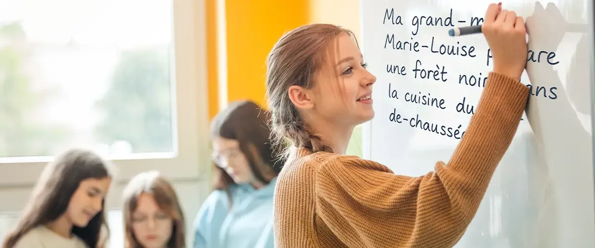 Une élève au tableau écrivant la phrase "Ma grand-mère Marie-Louise prépare une forèt noire dans la cuisine du rez-de-chaussée". Exercice sur les tirets - cours de français Acadomia