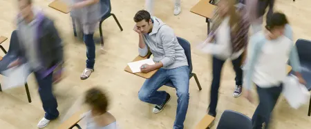 Un lycéen isolé et pensif reste assis à son bureau tandis que d'autres élèves s'agitent autour de lui dans une salle de classe