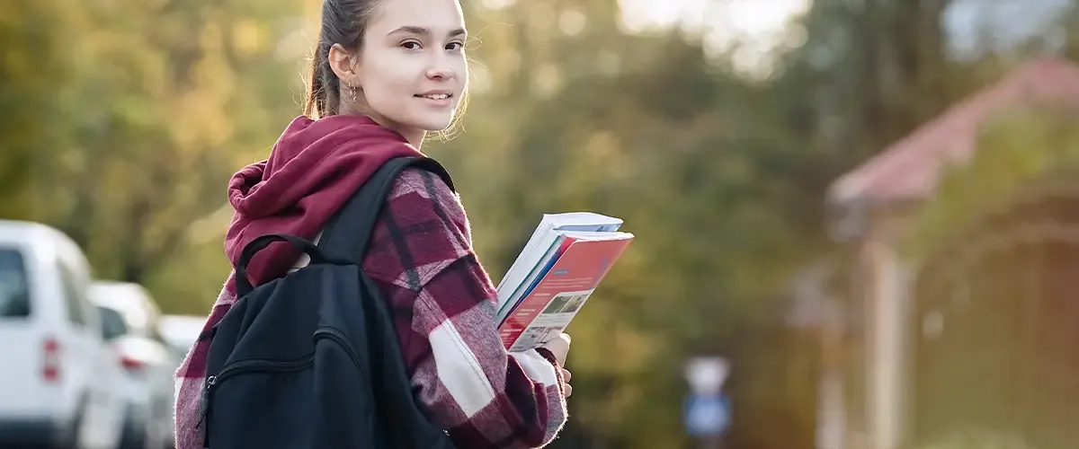 Une collégienne souriante portant un sac à dos et tenant des manuels scolaires marche dans une rue résidentielle
