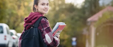 Une collégienne souriante portant un sac à dos et tenant des manuels scolaires marche dans une rue résidentielle