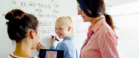 Deux collégiennes et une enseignante Acadomia travaillent ensemble sur des équations mathématiques devant un tableau blanc dans une salle de cours